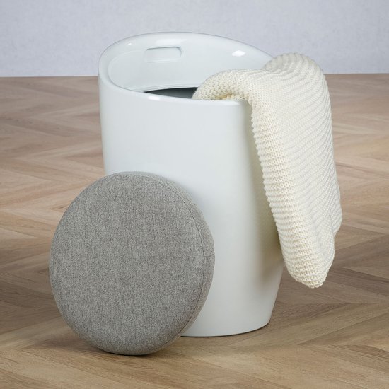 Witte cilindervormige opbergkruk met een los rond grijs zitkussen en een witte gebreide deken erin.