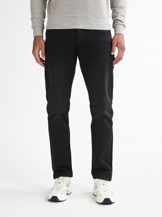 Man van voren gezien, draagt een grijze sweater en zwarte jeans met witte sneakers.