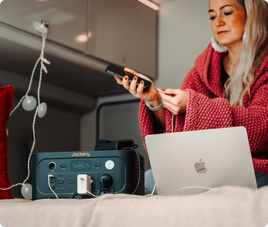 Donkergrijs Jackery Explorer 500 powerstation met oranje accenten, aangesloten op een telefoon en laptop, naast een vrouw met een koptelefoon.