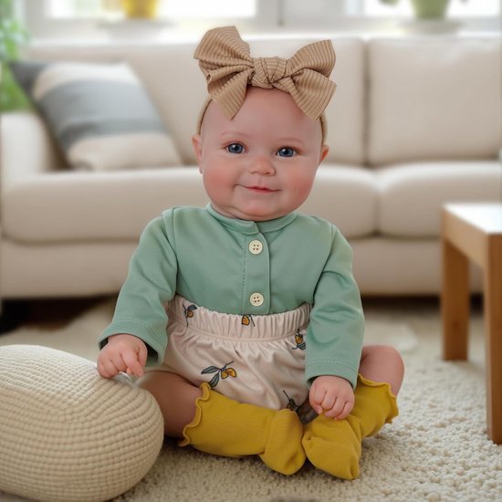 Baby met strik, groen shirt, luierbroekje met olijvenprint en gele sokjes, zittend op een kleed.