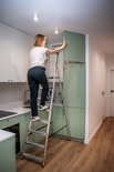 Een vrouw op een aluminium Baulich trap, reikend naar een kastdeur in een moderne keuken met mintgroene kasten.