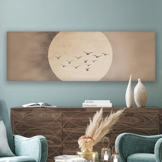 Langwerpig canvas met een grote, licht beige cirkel tegen een beige achtergrond. Een groep donkere vogels vliegt in formatie voor de cirkel. Hangt boven een donker houten dressoir met boeken en decoraties.