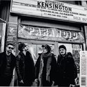 Vier bandleden staan buiten voor een bioscoopachtige aanplakzuil met de tekst 'KENSINGTON' en 'SOLD OUT'.