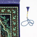 Tapis de prière bleu foncé avec une arche de mosquée et des motifs de feuilles verts/dorés, à côté d'un chapelet bleu avec un pendentif en forme de lune.