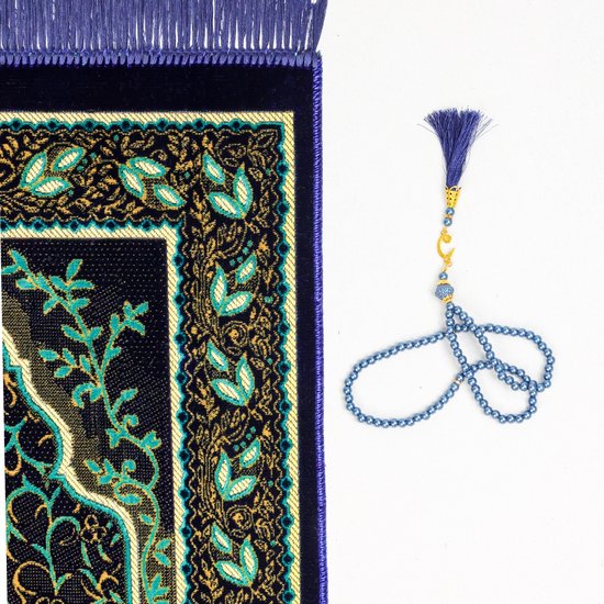 Tapis de prière bleu foncé avec une arche de mosquée et des motifs de feuilles verts/dorés, à côté d'un chapelet bleu avec un pendentif en forme de lune.