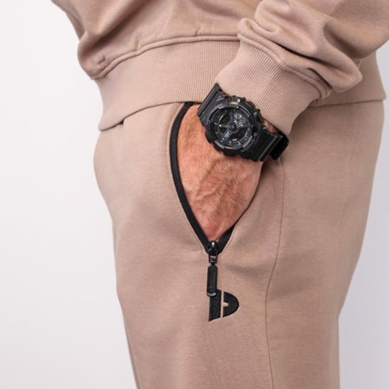Detail van een taupe-grijze joggingbroek met een rits en een klein logo, gedragen door een man.