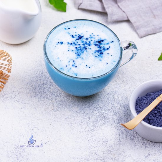 Glazen mok met blauwe matcha latte, bestrooid met matcha poeder. Naast een kannetje melk en een kommetje matcha.
