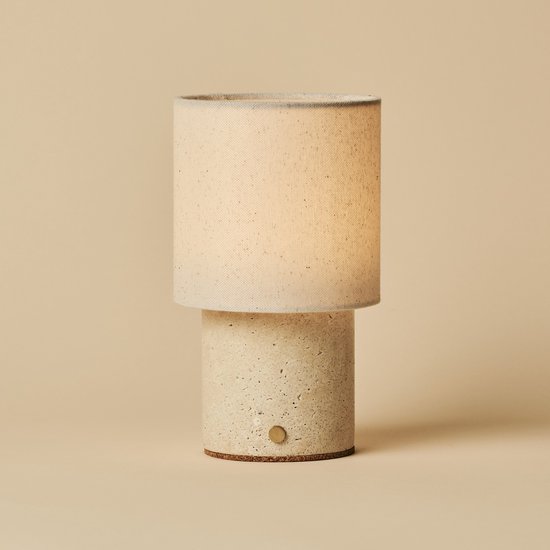 Cilindrische tafellamp met beige linnenkap en travertijnen voet, geïsoleerd op een beige achtergrond.