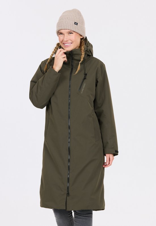 Model draagt een olijfgroene parka met rits. Ze heeft een beige muts op en haar blonde haar is in twee vlechten gevlochten. Ze houdt haar kin vast.
