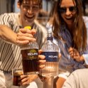 Twee flessen Mesamis: Caribbean Rum donkerbruin en London Juniper lichtroze. Mannen en vrouwen heffen hun glas.
