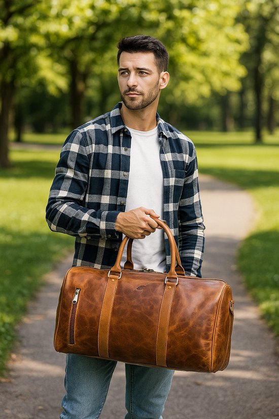Man met donker geblokt overhemd draagt een cognac leren weekendtas in een park.