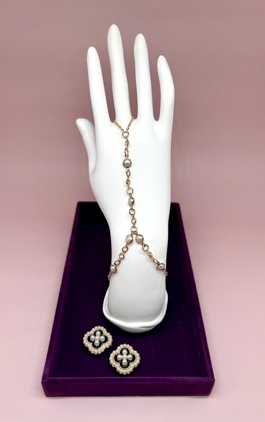 Gouden handketting met parels en zwarte klaveroorbellen op witte hand.
