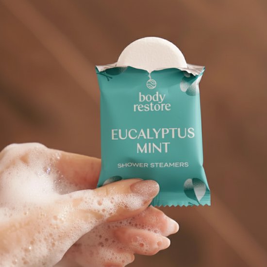 Hand houdt een verpakte Body Restore Eucalyptus Mint douchetablet vast met zeepbellen.
