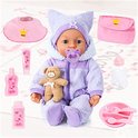 Een baby-pop met blauwe ogen en een paars, zacht pakje met muts. Rondom de pop liggen accessoires: een knuffelbeer, flesje, slabbetje, tasje, potjes en een bordje met bestek.
