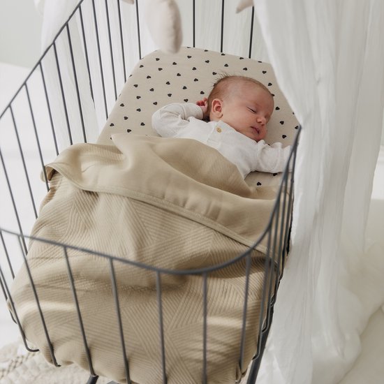 Slapende baby in een wieg, bedekt met een beige deken en een hoeslaken met hartjes.