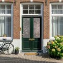Donkergroene voordeur met glasfolie met bloemen- en bladerpatroon, naast een fiets en struiken.