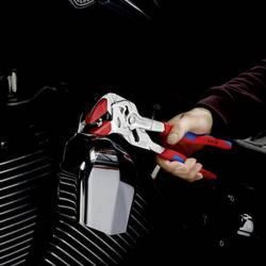 Hand houdt een tang met rode en blauwe handvatten vast, die een deel van een motorfiets aanpast.