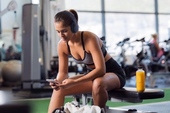 Vrouw in sportkleding zit op een bankje in de sportschool, kijkt op haar telefoon en draagt een donkerblauwe koptelefoon.