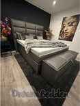 Grijze boxspring met hoog hoofdbord, witte matras, grijze deken, sierkussens en een opbergbank.