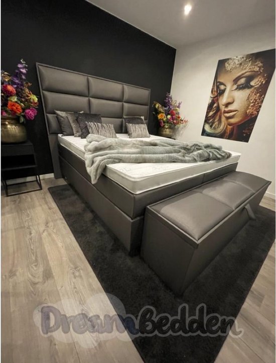 Grijze boxspring met hoog hoofdbord, witte matras, grijze deken, sierkussens en een opbergbank.