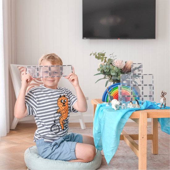 Een jongen met een gestreept shirt en blauwe shorts houdt twee sets Connetix-tegels voor zijn ogen, met een speeltafel met dieren en een regenboog op de achtergrond.