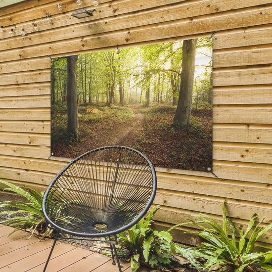 Tuinposter met een bos en een pad, hangend aan een houten wand met een zwarte stoel ervoor. De zon schijnt door de bomen.