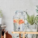 Glazen drankdispenser met 'Hello Summer' opdruk, gevuld met water en partjes grapefruit en takjes kruiden.