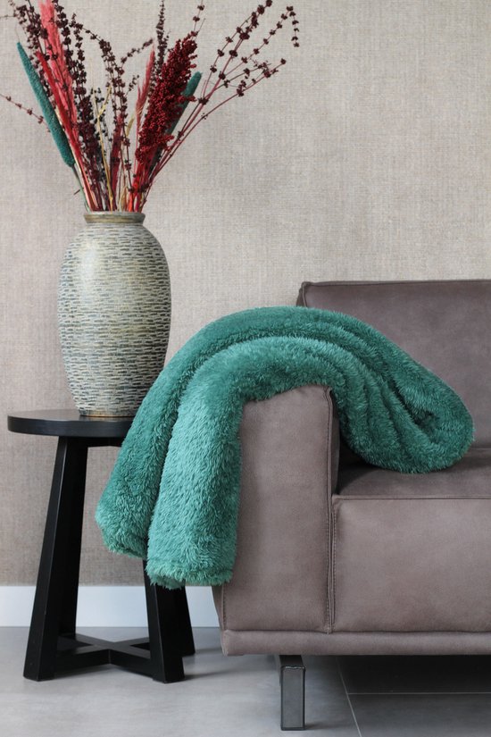 Teal fleece deken gedrapeerd over de armleuning van een grijze bank, naast een vaas met gedroogde bloemen.