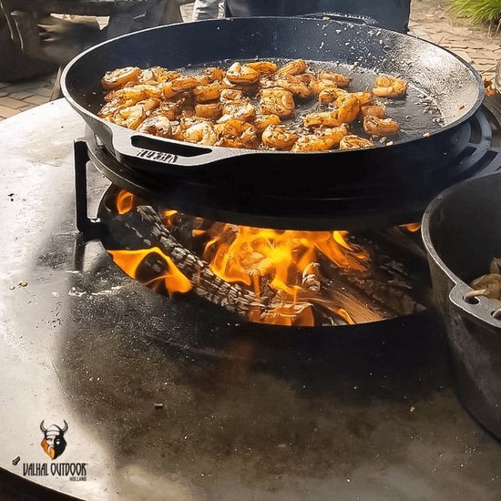 Grote gietijzeren pan met garnalen die worden bereid boven een open vuur op een metalen tafel.