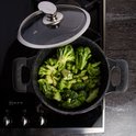 Zwarte pan met handvatten, gevuld met broccoli, staat op een gasfornuis met de glazen deksel ernaast.