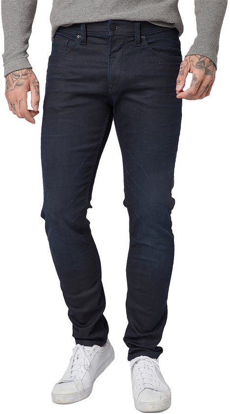 Een man met tatoeages draagt een grijze longsleeve en een donkerblauwe slim fit jeans.