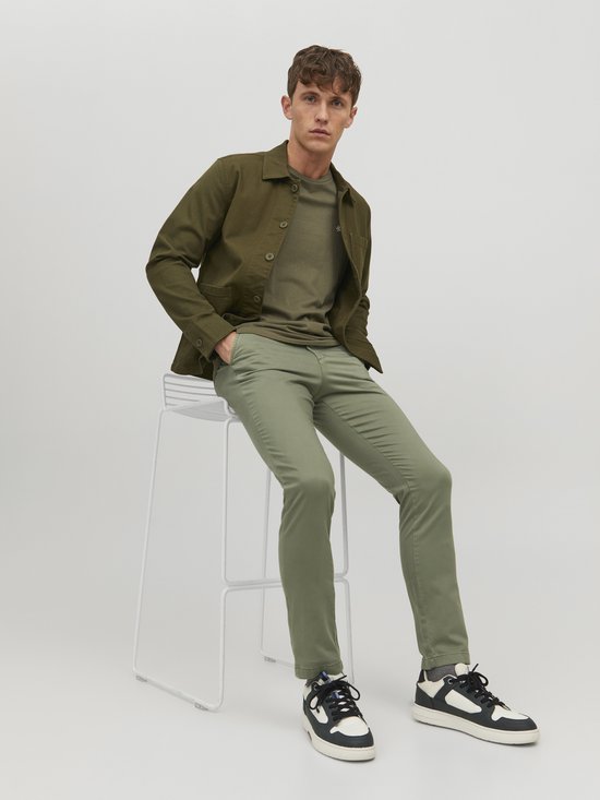 Man zittend op een barkruk, draagt olijfgroene chino broek met groen jack en t-shirt.