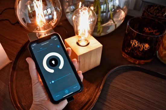 Hand houdt smartphone vast met Calex app, naast een glazen lamp en een houten lampvoet met gloeiende ledlamp.