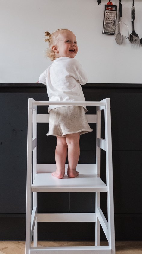 Kind met blonde knotjes in wit shirt en beige broek lacht op witte leertoren.