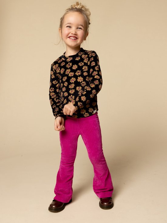 Een lachend meisje met blond haar in een knot, draagt een zwart bloemen t-shirt en roze corduroy broek.