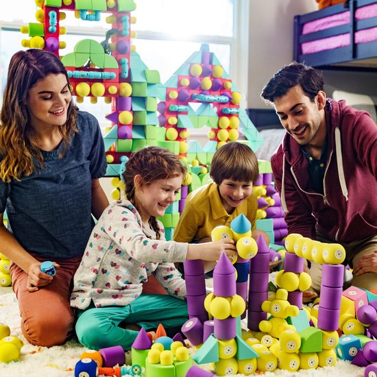 Een familie van vier speelt lachend op de vloer met een grote constructie van kleurrijke, zachte bouwblokken.