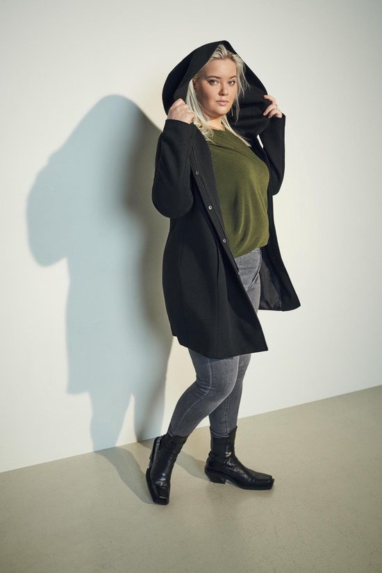 Vrouw met blonde haren, draagt een zwarte lange jas met capuchon, olijfgroene top en grijze skinny jeans.
