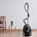 Zwarte stofzuiger met grijze slang en stang, staand op een houten vloer in een woonkamer.