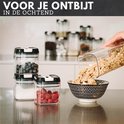 Doorzichtige voorraadpotten met zwarte deksels, gevuld met bosbessen, suiker en frambozen, terwijl muesli in een kom wordt gegoten.