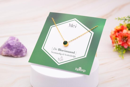 Gouden ketting met smaragdgroene hanger op een kaart met 'Mei - De Bloemmaand'.