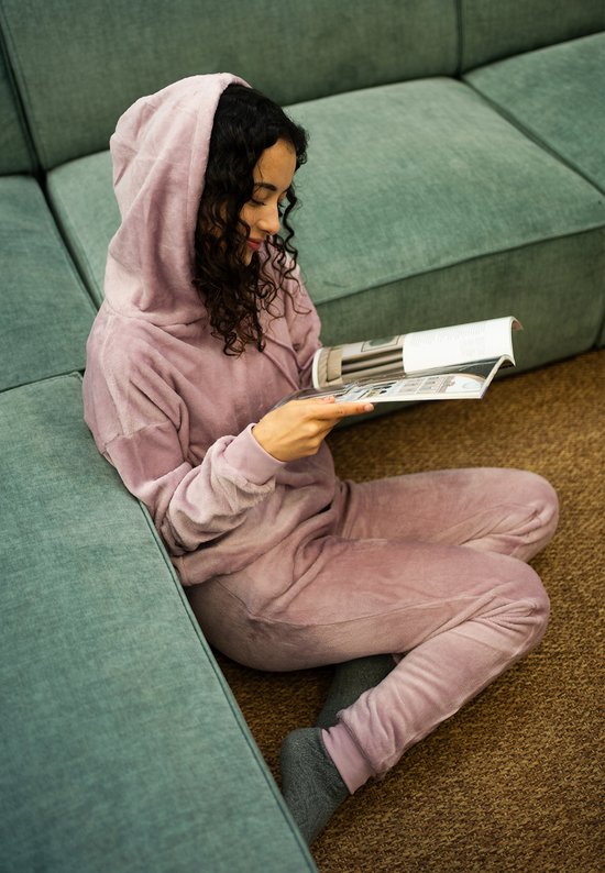 Vrouw met krullend haar leest een tijdschrift, zittend op de grond in een lila fleece loungepak.