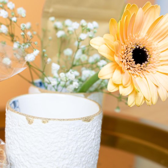 Witte mok met textuur en een Delfts blauw bloemenpatroon. Een oranje gerbera staat ernaast.