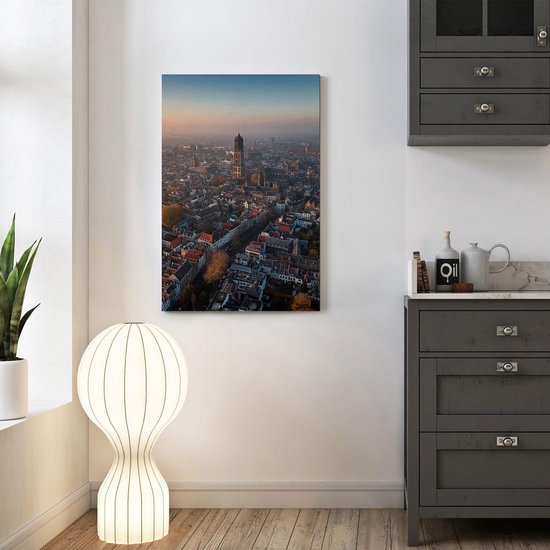 Une peinture de la skyline d'Utrecht au lever du soleil, avec la tour Domtoren bien visible. Le ciel est orange et bleu, et la ville est enveloppée de brume. Accrochée au-dessus d'une commode gris foncé.