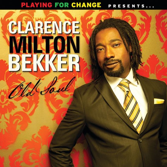 Clarence Milton Bekker met dreadlocks, baard, gestreept geel, bruin en wit gestreept, gele pochet, in een zwart pak.