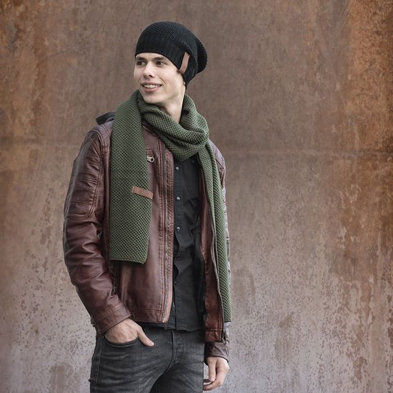 Jonge man met een zwarte beanie en een olijfgroene sjaal om zijn nek, draagt een bruine leren jas.