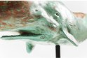 Close-up van een verweerd turquoise en bruin houten walvisbeeld, met een open mond en tanden, op een zwarte standaard.