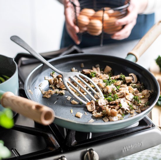 Frituren van champignons en kruiden in een koekenpan met houten handgreep op een fornuis. Een hand met een tang en een mand met eieren op de achtergrond.