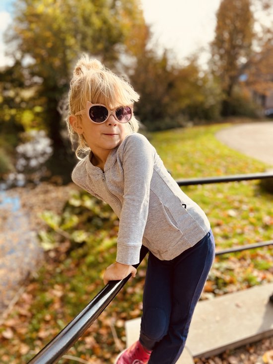 Meisje met blond haar in een grijze jas en kattenbril, leunend op een reling.