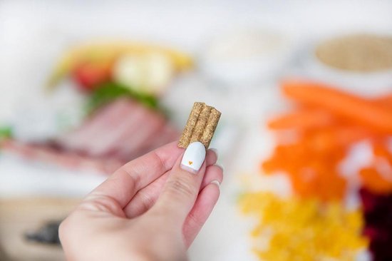 Hand met een hondenbrokje lam & rijst. Op de achtergrond ingrediënten.
