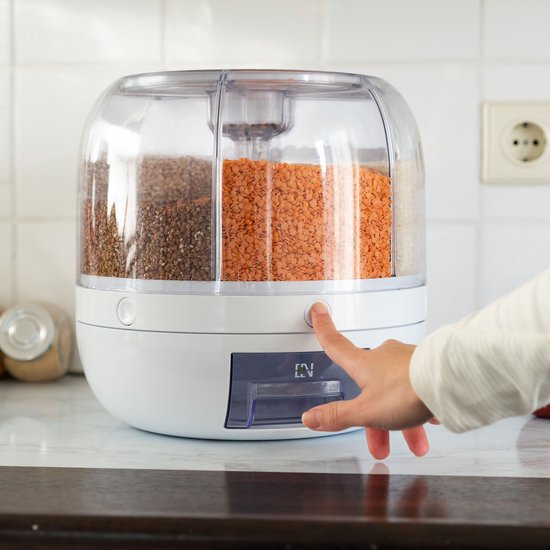 Een vinger drukt op de knop van een witte voorraaddispenser met vier vakken, gevuld met granen en linzen.